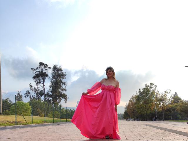 CelesteLincon in a pink, off-shoulder dress with flowing fabric, standing on a paved path, holding the skirt, smiling, with trees and a cloudy sky in the background.