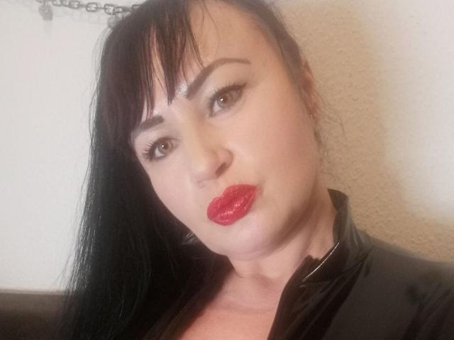 NanaGloss has pale skin, black hair with bangs, red glossy lips, and black eyeliner. She's wearing a black leather jacket, looking slightly pouty.