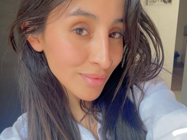 LeilaP tem a pele marrom média, olhos castanho escuros e um pequeno piercing no nariz. Ela está sorrindo, com os lábios rosados, vestindo uma camisa branca e tem o cabelo castanho escuro, molhado..