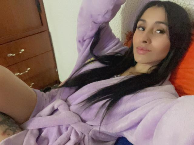 LeilaP, con el pelo largo y negro, vestida con una túnica morada, acostada en una cama, mirando hacia un lado con una ligera sonrisa, con la mano por encima de la cabeza, mostrando un tatuaje en el muslo..