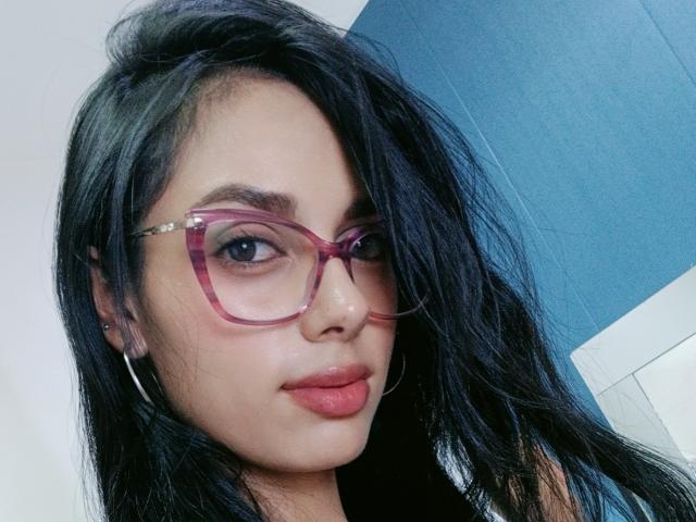 AnaisHilton with black hair, pink glasses, and pink lipstick, showing a cute smile with hoop earrings, wearing a black top, in a selfie.