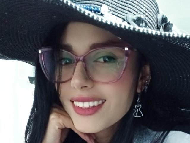 AnaisHilton with black hair, pink glasses, smiling, wearing a big black hat with white stripes, and silver earrings, showing white teeth and pink lips.