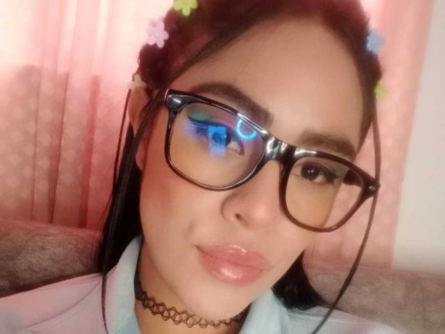 StellaBeckker with glossy pink lips, black choker, blue-green eyeshadow, black glasses, and flower headpiece in her dark hair, wearing a white shirt, looking at the camera.