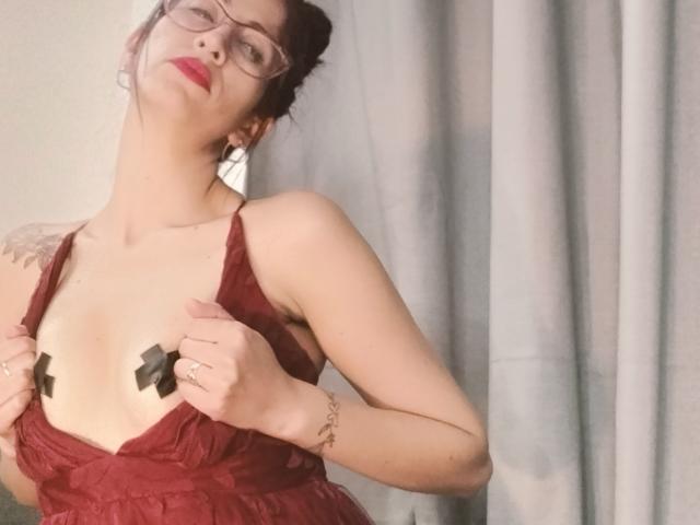 CateVanss with red lace dress, holding straps, exposing black tape on her tits, wearing glasses, red lipstick, tattoo on left shoulder, gray curtain background.