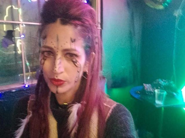 CateVanss mit roten Haaren, weißer Gesichtsfarbe, schwarzen Punkten, rotem Lippenstift und einem schwarzen Nasenring, trägt ein graues Oberteil und eine beige Weste, in einem grün beleuchteten Raum..