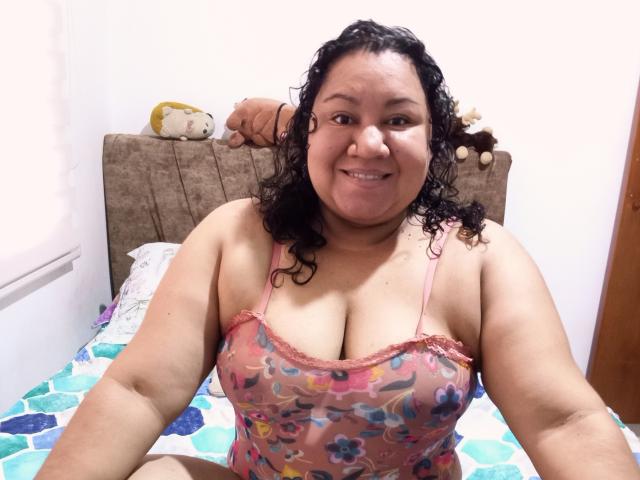 Curvy HiyunaMei with dark curly hair, smiling, wearing a pink floral tank top, showing her large boobs, sitting on a bed with colorful patterned sheets.