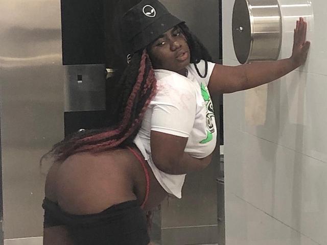 Dilay with dark skin, long red and black braids, wearing a black cap, white shirt, and black shorts pulled down, exposing her big ass, leaning on a white wall.