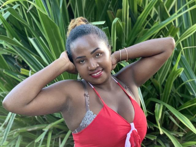 NenetteFun with dark skin, curly brown hair in a bun, smiling, red top showing tits, hands behind head, green plant background, red lipstick, and a bracelet on her right wrist.