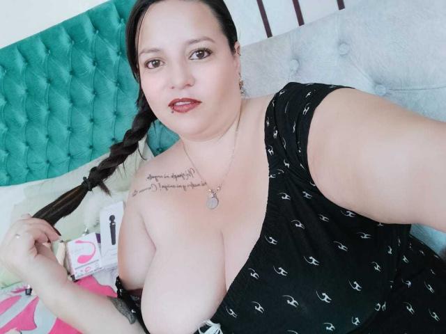 SkayLaraWalker, avec de grandes seins, vêtue d'une robe noire, les cheveux tressés, un tête de lit vert, un tatouage sur l'épaule droite, un collier, souriante, peau pâle, rouge à lèvres rouge, allongée sur un lit..