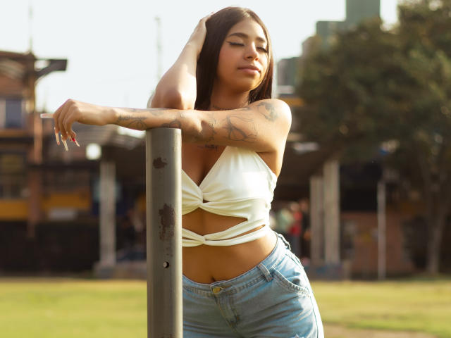 LiaAnahiz with tan skin, tattoos on her arm, brown hair, white tied-up crop top, showing her tits, and blue jeans, leaning on a pole, eyes closed, in sunlight.