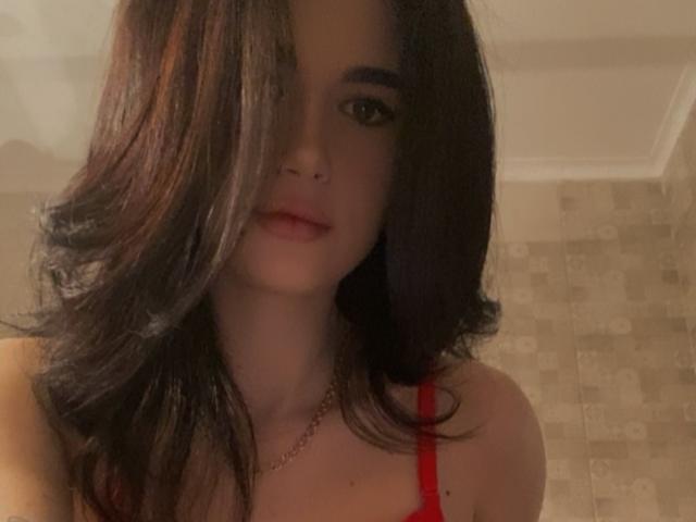 TiffanyNannette, con cabello oscuro y ondulado que cubre un ojo, vistiendo un sostén rojo, mostrando sus senos, y un aro nasal discreto, mirando a la cámara con una ligera sonrisa..