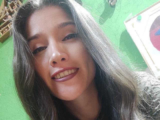 AliceMiz has long brown hair, brown eyes, and a small smile with burgundy lipstick. She's looking down at the camera, showing her nose and teeth. Green wall background.