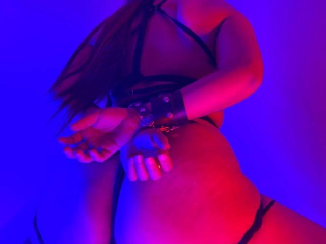 AbbySton in zwarte, open lingerie, polsen vastgebonden, handen voor haar, roze en blauwe verlichting, lang bruin haar, zichtbare billen, ring aan haar vinger, rode en roze highlights..
