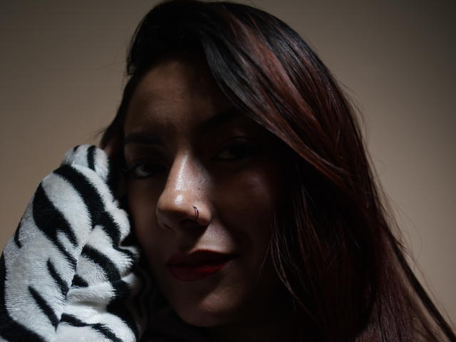 AbbySton hat dunkelbraunes Haar mit roten Strähnen, einen kleinen Nasenring und roten Lippenstift. Sie trägt ein zebra-gestreiftes Hemd und blickt mit einem leichten Lächeln nach unten..