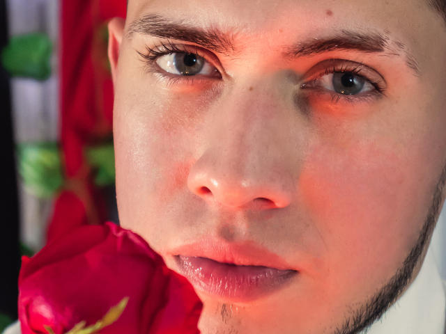 SebastiianMaxime's face, with red rose near nose, showing subtle pink blush, thin eyebrows, and slightly parted lips, has a soft, slightly shiny complexion. His eyes are green.