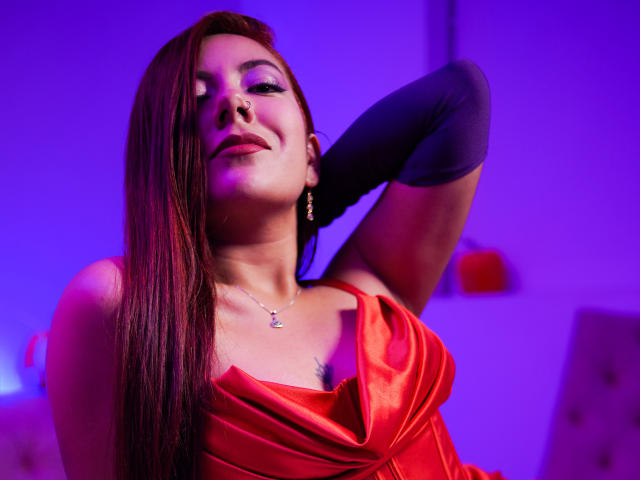 AbbySton, avec de longs cheveux bruns, une robe rouge, des manches noires, un anneau au nez et un collier, posant les bras derrière la tête, éclairage violet et rouge, léger sourire, et décolleté visible..