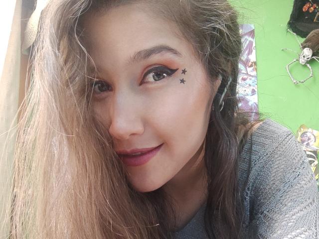 AliceMiz with long, messy brown hair, black star tattoos under her right eye, gray sweater, and pink lipstick, smiling at the camera. Her ass is not visible.