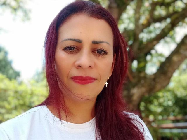 SashaMissHot with red hair, white shirt, cross earrings, and red lipstick, showing a slightly annoyed or confused expression. Her tits are average and her pussy is not visible.