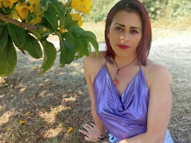 SashaMissHot with red-dyed bob, purple halter top, showing her small tits, red lipstick, necklace with pink gem, sitting under yellow flowers, grassy ground.