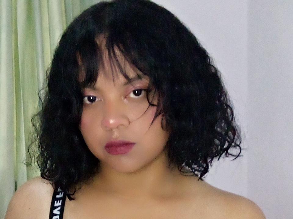 NicolTaylorX ha la pelle marrone media, i capelli neri e ricci che le arrivano alle spalle, e guarda direttamente la fotocamera con il rossetto rosso e un'espressione neutra. Indossa un reggiseno nero..