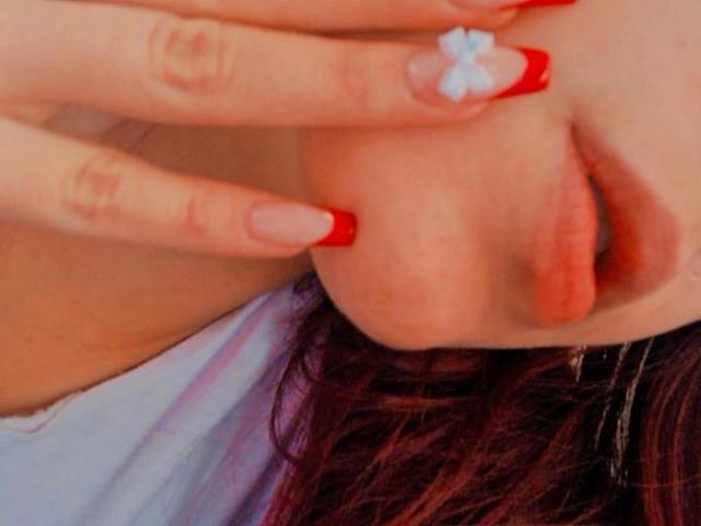 De lippen van Linsay Colemann zijn lichtjes open, en haar rood gelakte vingers raken haar wang. Ze heeft een blauwe bloem op haar nagel en roze uiteinden aan haar nagels. Haar paarse haar is zichtbaar..