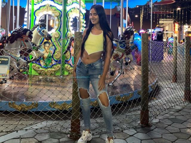 TamaraPeach stands in front of a carousel, wearing a yellow crop top, ripped blue jeans, and white sneakers, with long black hair, smiling, and showing her toned midriff.