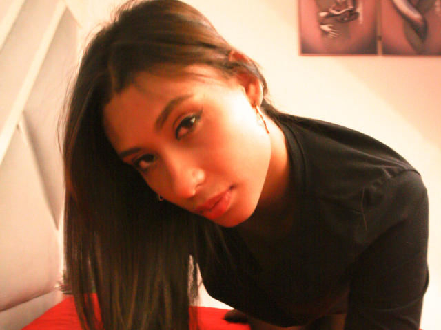 VanessaLove, with long brown hair, leaning forward, wearing a black top, has a cute, slightly mischievous look, with subtle makeup and a slight pout. Soft, warm lighting.