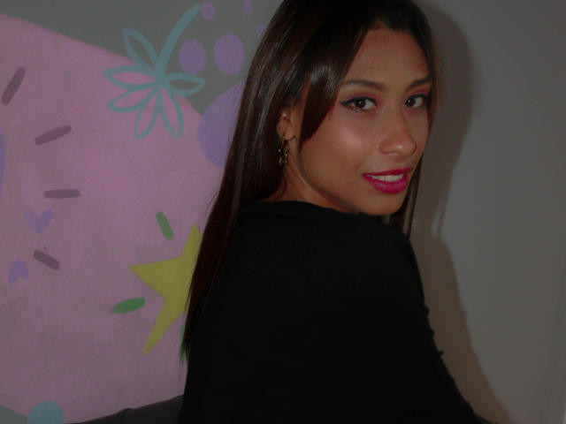 VanessaLove with dark brown hair, pink eyeliner, and pink lipstick, wearing a black top, smiling at the camera, showing her ass slightly. She's looking over her shoulder.