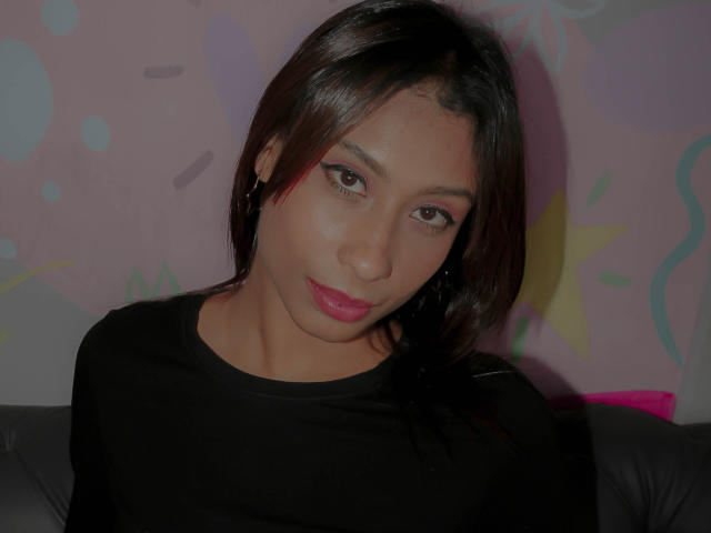 VanessaLove has medium brown skin, black straight hair, pink lipstick, black top, and brown eyes with winged eyeliner, sitting against a pink, patterned wall.