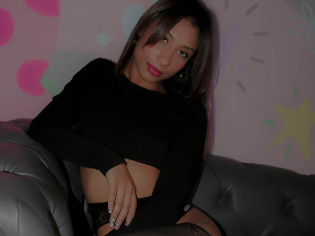 VanessaLove with brown hair, pink lipstick, black long-sleeve top, exposing her midriff, sitting on a gray leather couch, wearing black lace panties, looking at the camera.