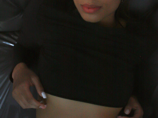 VanessaLove's upper body, wearing a black shirt, with her right hand pulling it up to reveal her flat, tan midriff and slightly parted red lips. Her nails are white.
