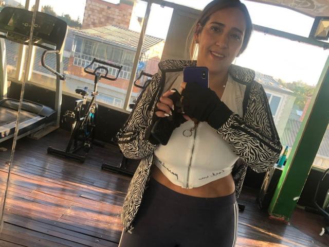 BlueWaters with dark hair, wearing a zebra-patterned jacket, white sports bra showing her tits, black gloves, and navy pants, taking a selfie in a gym.