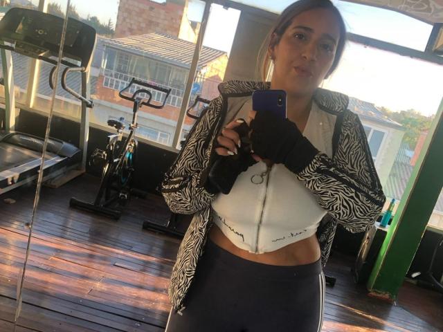 Mulher de pele clara, com seios médios, vestindo um top de ginástica branco com zíper, luvas pretas e um moletom com estampa de zebra, tirando uma selfie no espelho em uma academia..