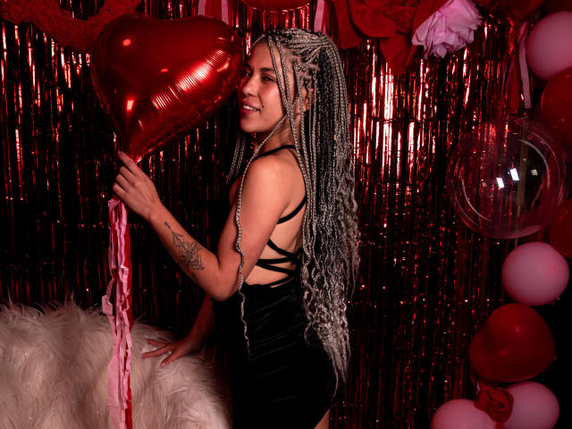 HellenK with long silver braids, black velvet dress, holding red heart balloon, tattoo on arm, smiling, standing next to white furry pillow, red and white balloons around.