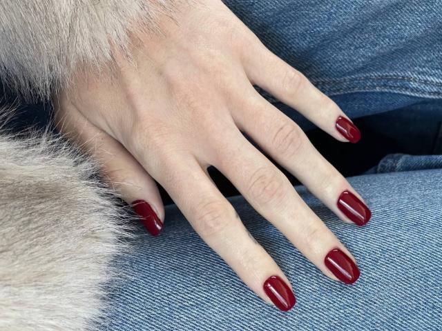 Tsukerberg's hand with shiny red nail polish rests on blue denim, her fingers slightly spread, showing off neatly trimmed, pale skin and a fluffy grey fur sleeve.