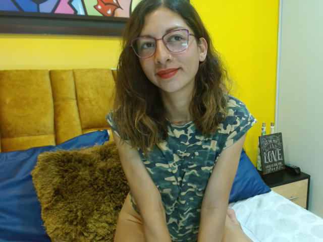 VioletSingh, met golvend bruin haar, een bril en rode lippenstift, draagt een camouflagehemd en zit op een bed met een geel hoofdeinde en een bruin kussen. Haar achterkant is niet zichtbaar..