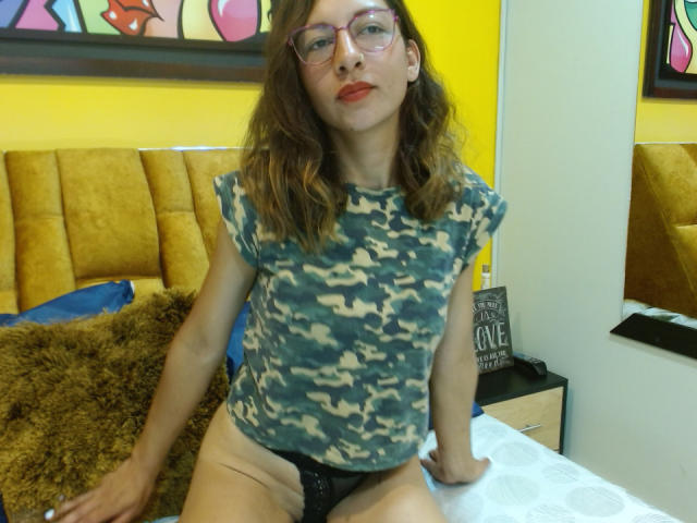 VioletSingh, met golvend bruin haar, een bril en rode lippenstift, draagt een camouflage top en zwarte kanten onderbroek, en zit op een bed met een geel hoofdeinde en een bruin kussen..