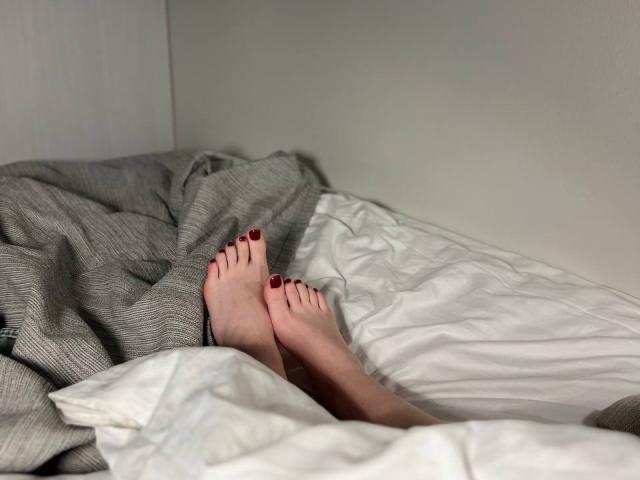 MiaMoone's bare feet with red-painted toes are crossed on a bed with gray and white sheets.