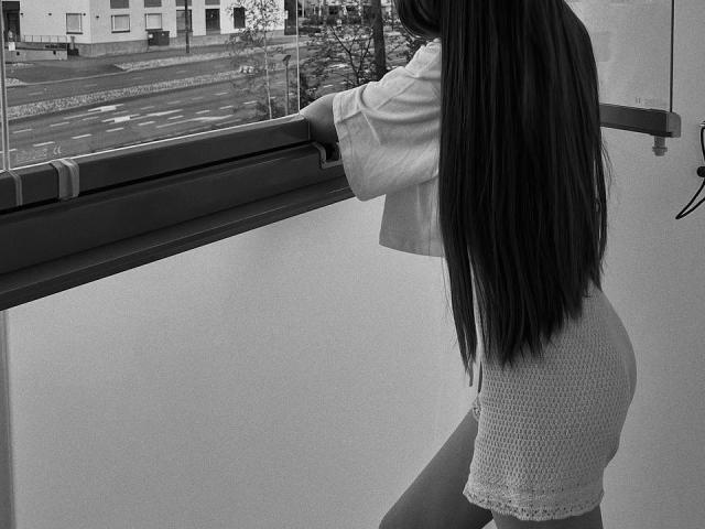 MiaMoone, con lunghi capelli neri e lisci, indossa una camicia larga e dei pantaloncini aderenti con bordi in pizzo, in piedi vicino alla ringhiera di un balcone, che guarda fuori, in una fotografia in bianco e nero..
