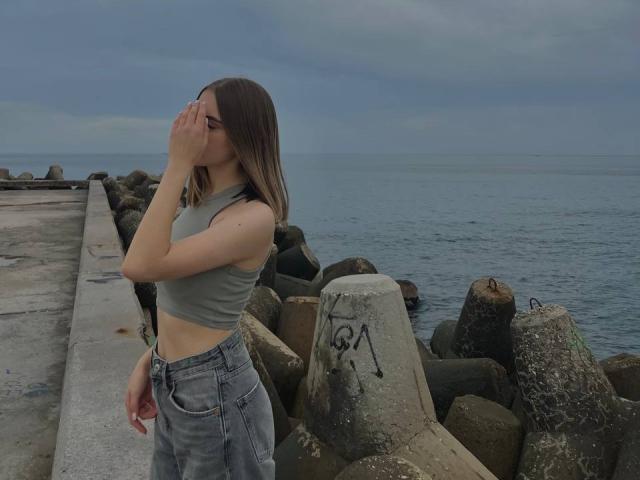 MiaMoone, con i capelli castano di lunghezza media, indossa un top corto verde e jeans blu, è in piedi vicino all'oceano, con una mano sulla testa, guarda di lato, mostrando il sedere..