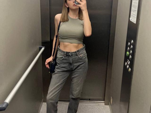 MiaMoone in un ascensore, indossa un top corto verde che mette in mostra i suoi piccoli seni e dei jeans grigi a vita alta, leggermente larghi. Tiene in mano un telefono e ha una borsa nera..