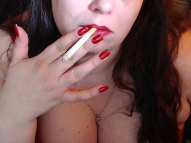 MayaSmith with red nail polish, holding a lit cigarette to her red lips, her hand near her tits, wearing a necklace with a small cross, dark wavy hair.