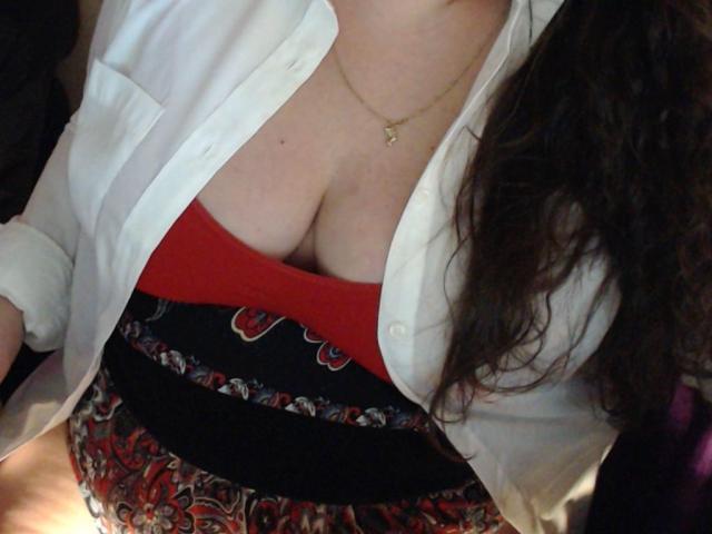 MayaSmith with large, rounded boobs, wearing a red top, black patterned skirt, and white open shirt, gold necklace, wavy brown hair, visible cleavage, and a cute gold pendant.