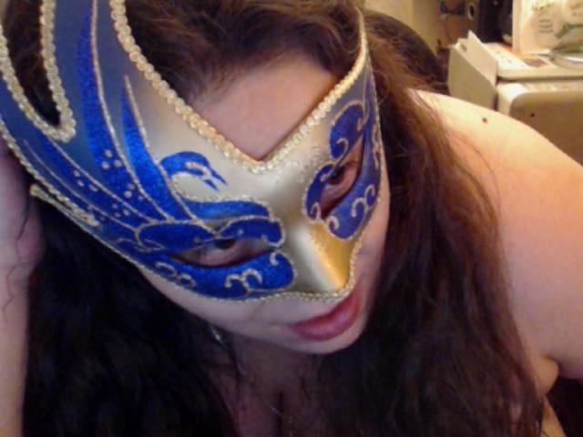 MayaSmith with long brown hair, wearing a blue and gold masquerade mask, leaning forward, showing her red lips and partially visible bare tits, in a dimly lit room.