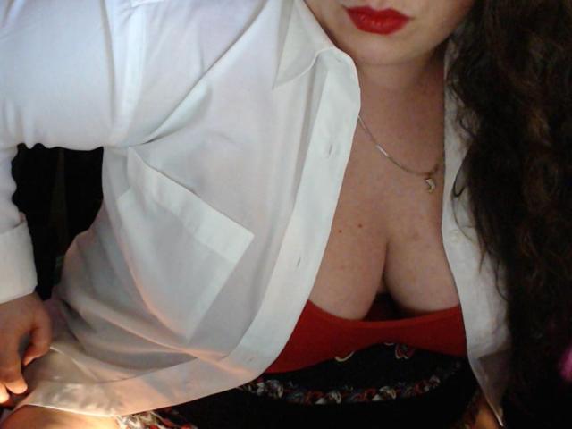 MayaSmith with red lipstick, white shirt, and deep cleavage in a red top, hands on her hip, wearing a patterned skirt, necklace, and long curly brown hair.