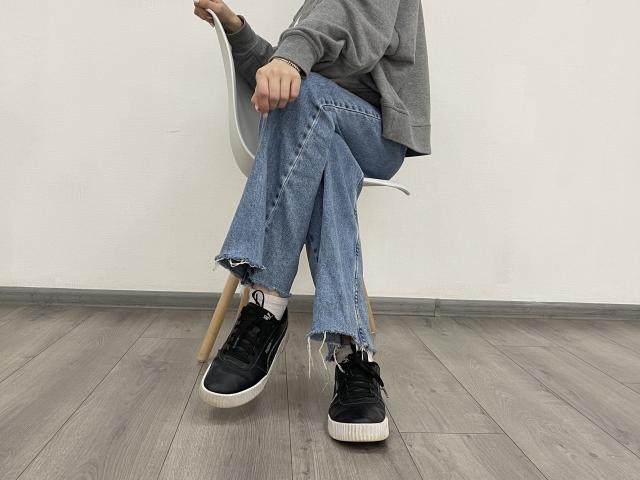 MiaMoone sitting on a chair, wearing a grey sweatshirt, blue frayed jeans, and black sneakers with white soles, on a wooden floor.