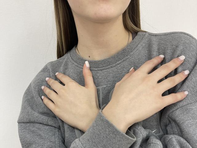 Le mani di MiaMoone, con le unghie dipinte di bianco, sono distese e coprono il suo petto. Indossa una camicia grigia a maniche lunghe. I suoi capelli castani sono sciolti, e il suo viso è parzialmente visibile..