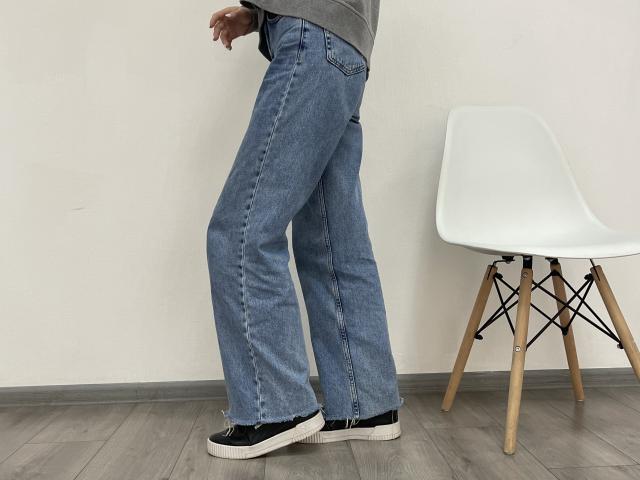 MiaMoone's ass in blue, flared, slightly frayed jeans, black and white sneakers, gray top, standing next to a white, wooden-leg chair on gray wood floor.