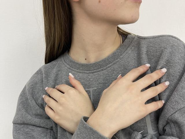 MiaMoone con le mani sul petto, felpa grigia, capelli castani lunghi, pelle chiara, smalto bianco sulle unghie, piccola macchia sulla nuca, leggero profilo, illuminazione soffusa, posa informale..