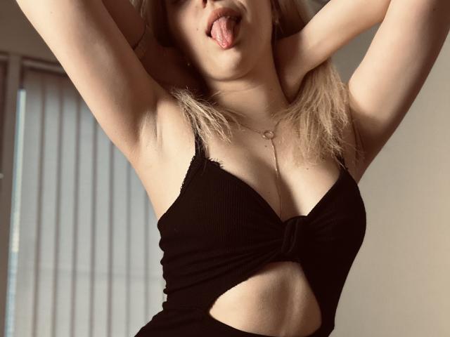 SugarrX with arms raised, tongue out, wearing a black cut-out top showing her tits and midriff, blonde hair, light skin, small gold necklace.